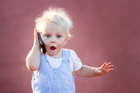 Cute baby boy playing with mobile phone in the park, digital technologies in the hands of a child. Portrait of toddler with smartphoneの写真素材