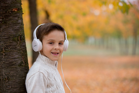 Sweet portrait of a preschool child, listening music with headphones and mobile phone in autumn park on sunsetの写真素材