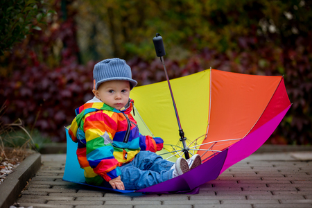 Sweet funny child with rainboy coat and multicolored umbrella jumping on puddles iand playing outdoors after rainの写真素材