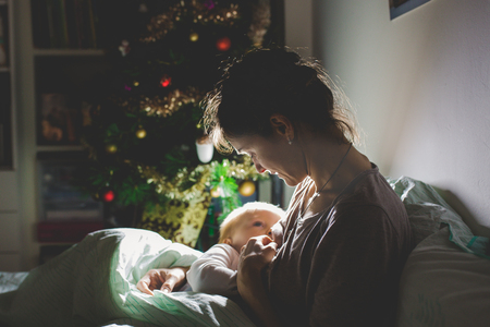 Young beautiful mother, breastfeeding her newborn baby boy at night, dim light. Mom breastfeeding toddler on Christmasの写真素材