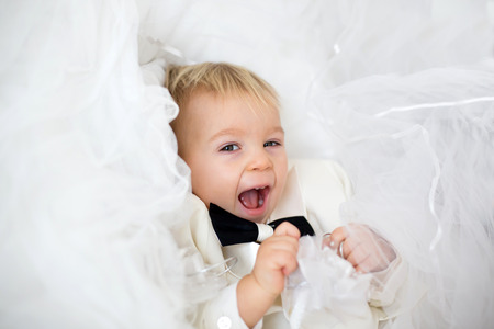 Portrait of elegant handsome little boy in a tuxedo, lying in bed on his moms bridal dress, ready to go to weddingの写真素材