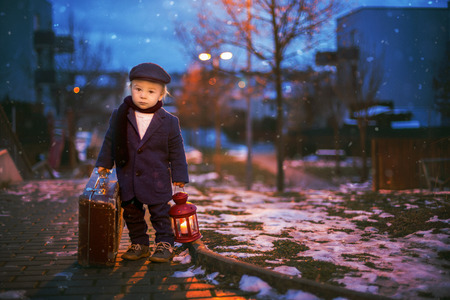 Toddler boy, standing on stairs, holding lantern and old suitcase, street view of Prague behind him, snowy eveningの写真素材