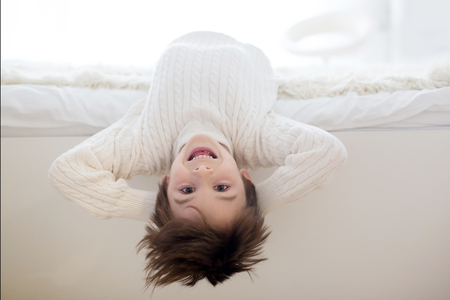 Cute portrait of child upside down, lying on bed, smiling at camera and playing with toyの写真素材