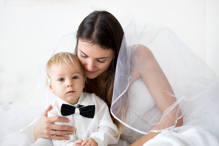 Portrait of elegant handsome little boyand mom as bride, lying in bedon her wedding day, dressed in bridal dress, baby with bowの写真素材