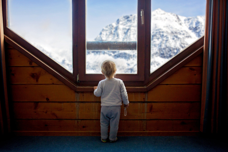 Toddler child standing in front of a big french doors, leaning against it looking outside at a snowy nature, playing with toysの写真素材