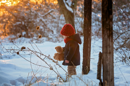 Baby playing with teddy in the snow, winter time. Little toddler boy in blue coat, holding teddy bear on sunset, playing outdoors in winter park. Children play in snowy parkの写真素材