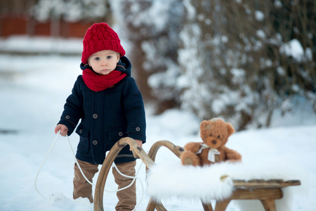 Baby playing with teddy in the snow, winter time. Little toddler boy in blue coat, sliding on sledge, holding teddy bear, playing outdoors in winter park. Children play in snowy parkの写真素材