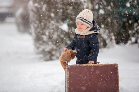 Baby playing with teddy in the snow, winter time. Little toddler boy in blue coat, holding suitcase and teddy bear, playing outdoors in winter park. Children play in snowy parkの写真素材