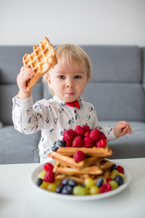 Sweet toddler birthday boy, eating belgian waffle with raspberries, blueberries, coconut and chocolate at homeの写真素材