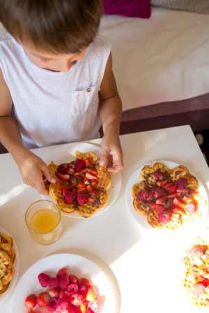 Sweet birthday boy, eating belgian waffle with strawberries, raspberries and chocolate at homeの写真素材