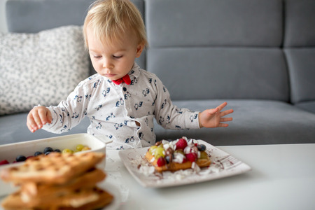 Sweet toddler birthday boy, eating belgian waffle with raspberries, blueberries, cocnut and chocolate at homeの写真素材
