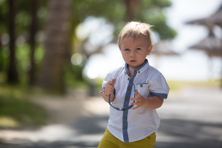 Adorable fashion baby toddler boy, dressed in casual clothes, walking in a beautiful resort gardens on the island of Mauritiusの写真素材