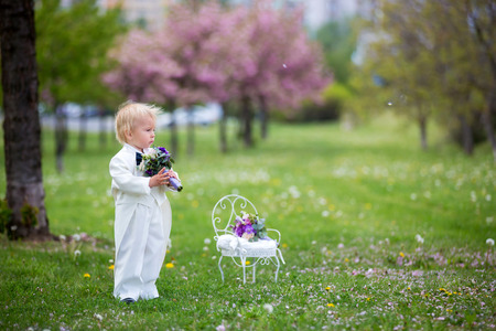 Beautiful toddler boy, dressed in white tuxedo, holding gorgeous flower bouquet for mothers day, smiling happily and giving it to mom in parkの写真素材