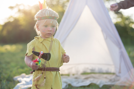 Cute boy dressed in traditional  native american costume playing with hatchet outdoorsの写真素材