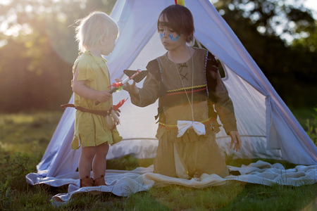 Cute boys dressed in traditional native american costume while playing outdoors in the park with hatchetの写真素材