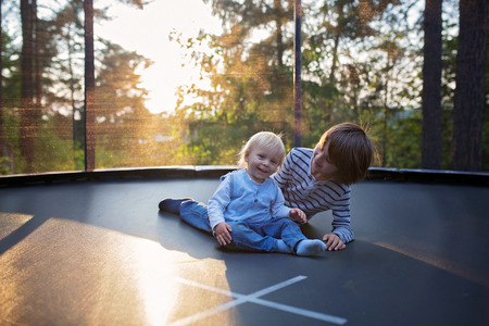 Sweet preteen boy and his toddler brother jumping on trampoline. Happy children jumping on sunset, making different shapesの写真素材