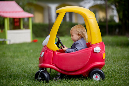 Cute toddler boy, riding big plastic red car toy in the park, springtime