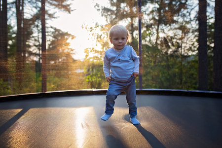 Sweet toddler baby boy jumping on trampoline. Happy little blond child jumping on sunset for the first time and having fun outdoorの写真素材