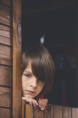 Preteen boy in red sweatshirt, hiding behind a wooden door, looking scared and sad