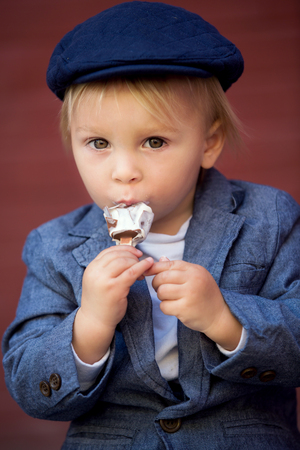 Cute child, boy in vintage cloths, eating lollipop ice cream, sitting on vintage suitcase in front of a red brick wall, summertimeの写真素材