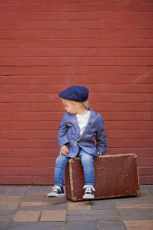 Cute little toddler child, sitting on a vintage suitcase in front of red bricks wall, dressed smart casual, summertimeの写真素材