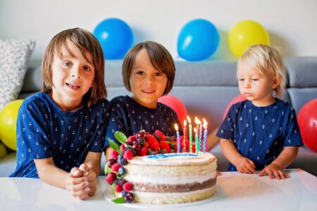 Beautiful eight years old boy and his brothers in blue shirts, celebrating his birthday, blowing candles on homemade baked cake, indoors. Birthday party for kidsの写真素材