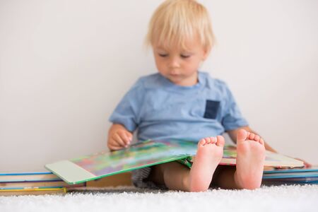 Sweet toddler boy, reading book at home, sitting on the ground, lots of books around himの写真素材
