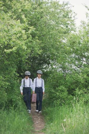 Sweet children in vintage clothing, hat, suspenders and white shirts, holding suitcase, running in...