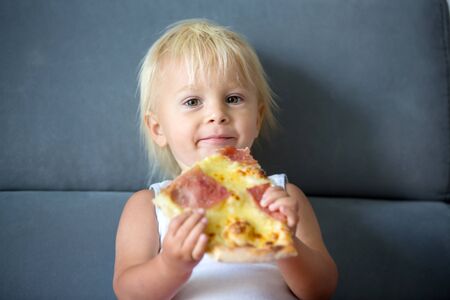 Cute little Caucasian kid eating pizza. Hungry child taking a bite from pizza on a pizza party. Smeared toddlerの写真素材