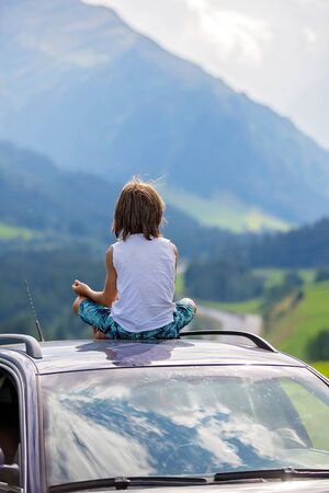 Cute child, boy sitting on a car ceiling, meditating enjoying the quietness of nature, travel on the road to scenic mountains, summertimeの写真素材