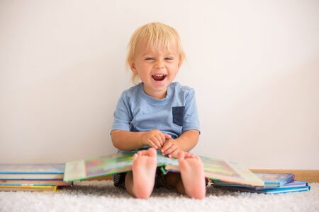 Sweet toddler boy, reading book at home, sitting on the ground, lots of books around himの写真素材