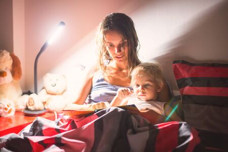 Mother, reading a book to her toddler boy at home at night, night stand lamp turned on, mom and baby...