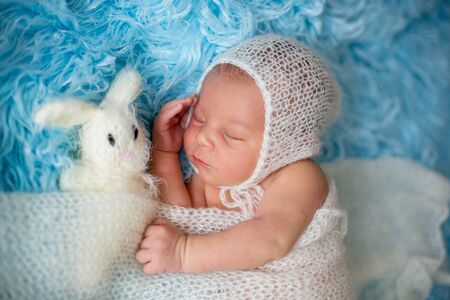 Little cute newborn baby boy, sleeping wrapped in white wrap, holding little knitted toyの写真素材