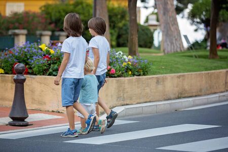 Three children, boys, brothers, holding hands and crossing a street on a striped crosswalk, checking for cars before, safety crossing the street summertimeの写真素材