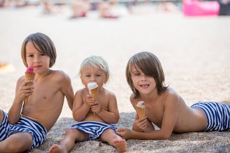 Sweet blonde toddler boy and his to older brothers, eaiting ice cream on the beach, summertime