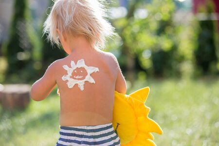 Toddler child with suntan lotion shaped as sun on his back, going at the beach with toys and flufy sun toy, walking outdoors back to cameraの写真素材