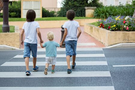 Three children, boys, brothers, holding hands and crossing a street on a striped crosswalk, checking for cars before, safety crossing the street summertimeの写真素材