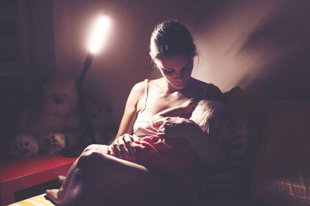 Mother, breastfeeding her toddler boy at home at night, night stand lamp turned on, mom and baby huggingの写真素材