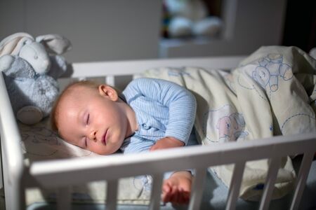 Adorable newborn baby boy, sleeping in crib at night. Little boy in white pajamas taking a nap in dark room. Bedroom interi?r with lamp and teddy bear in the backgroundの写真素材