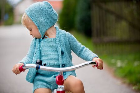 Adorable toddler boy with knitted outfit, riding tricycle on a quiet village street, summertimeの写真素材