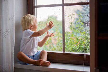 Adorable little boy, sitting on window shield, looking through the window on a rainy day, summertimeの写真素材