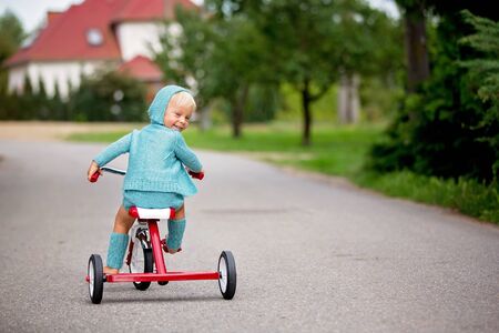 Adorable toddler boy with knitted outfit, riding tricycle on a quiet village street, summertimeの写真素材