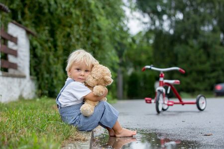 Cute little boy, with teddy bear toy, sitting on the street in the rain, barefeet, his tricycle on a sideの写真素材