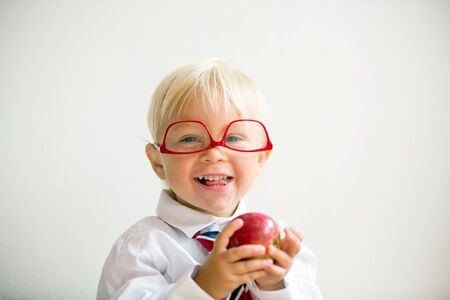 Cute kid, wearing glasses and eating apple, dressed for first day of school or kindergarden, happy smilingの写真素材