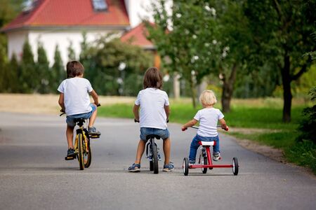 Three children, brothers, riding bike, tricycle and balance bike on the street in quiet village, happily competingの写真素材