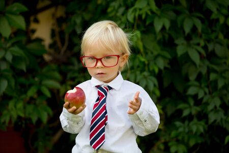 Cute kid, wearing glasses and eating apple, dressed for first day of school or kindergarden, happy smilingの写真素材