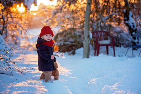 Baby playing with teddy in the snow, winter time. Little toddler boy in blue coat, holding teddy bear on sunset, playing outdoors in winter park. Children play in snowy parkの写真素材