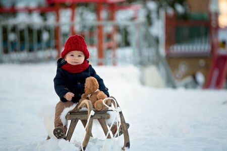 Baby playing with teddy in the snow, winter time. Little toddler boy in blue coat, sliding on...