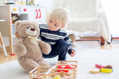 Sweet toddler boy, playing with teddy bears and wooden toys from kids kitchen set on the floor at homeの写真素材