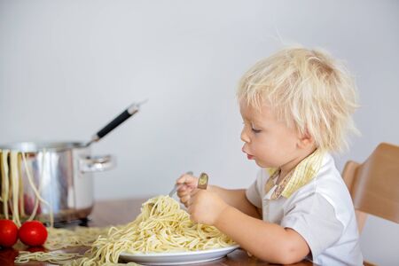 Little baby boy, toddler child, eating spaghetti for lunch and making a mess at home, pot with spaghetti and tomatoes on the tableの写真素材